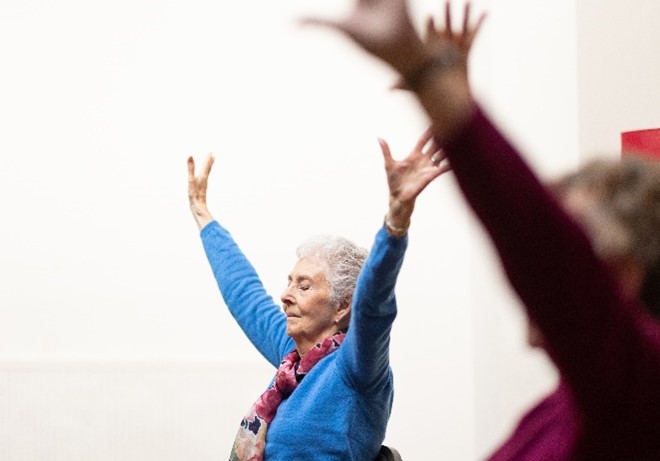 An older woman wearing blue shirt with pink scarf in pose with arms raised and eyes closed